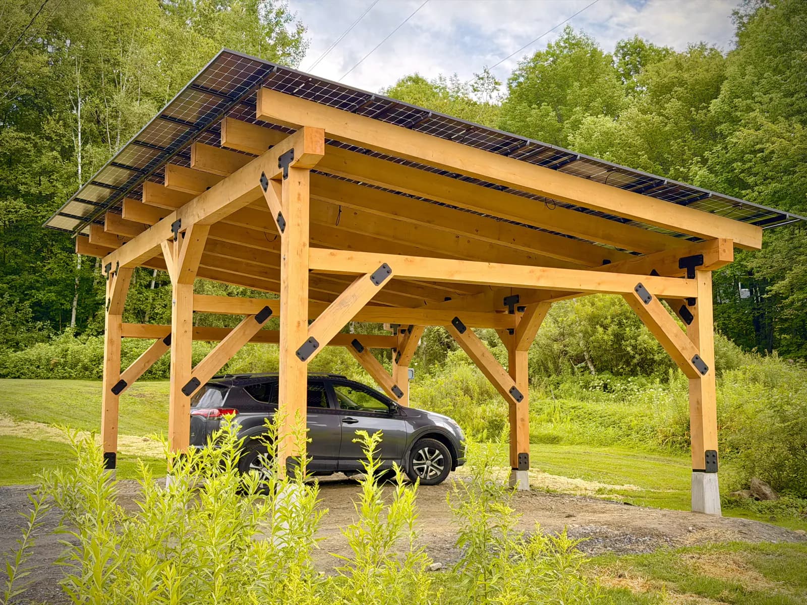 Completed timber-frame Sunport with a solar roof sheltering a parked car at the edge of a Vermont lawn