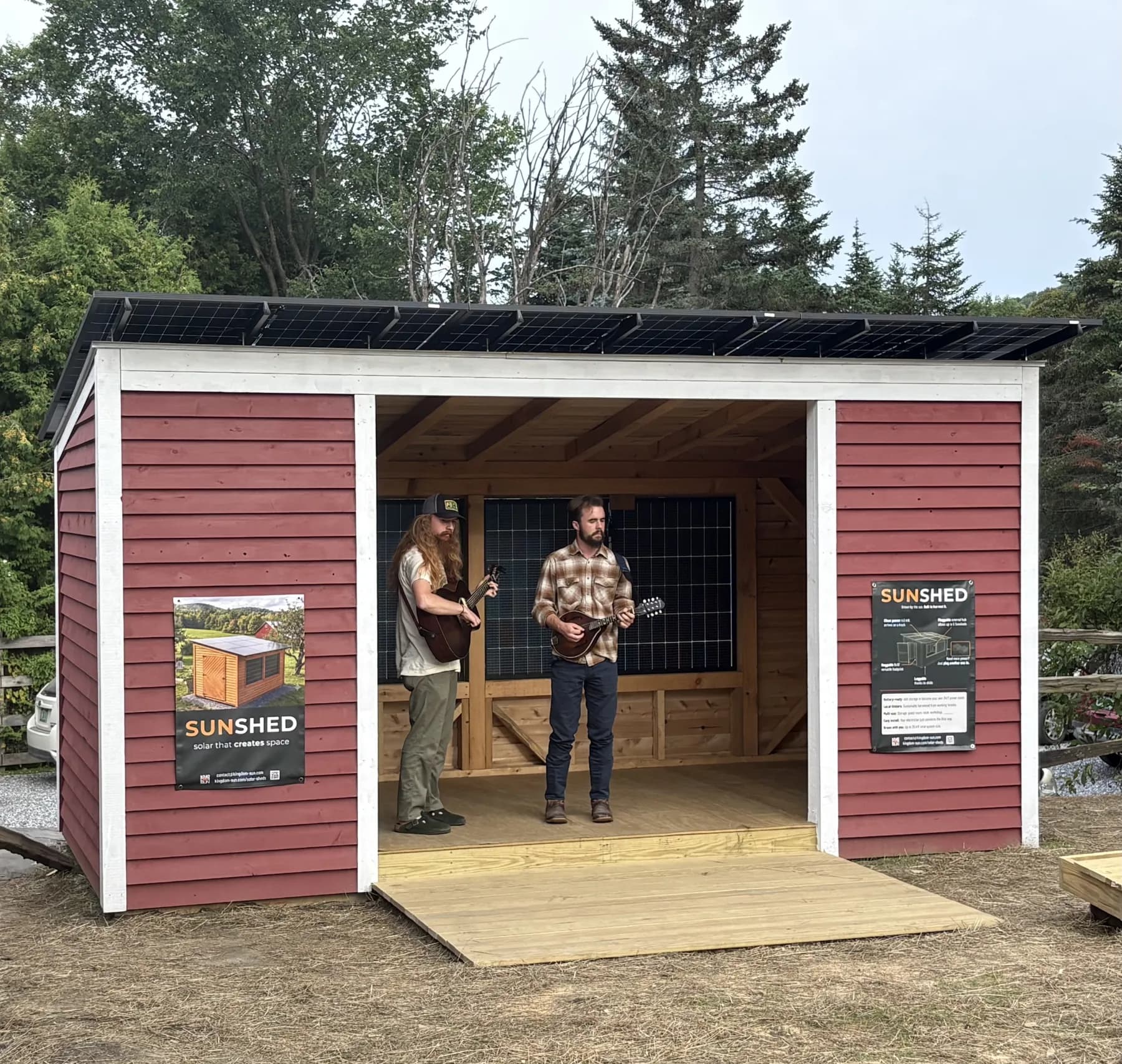 Two musicians playing guitar and mandolin inside a barn-red Sunshed set up as a stage at Parker Pie in West Glover, Vermont, solar panels along its roof